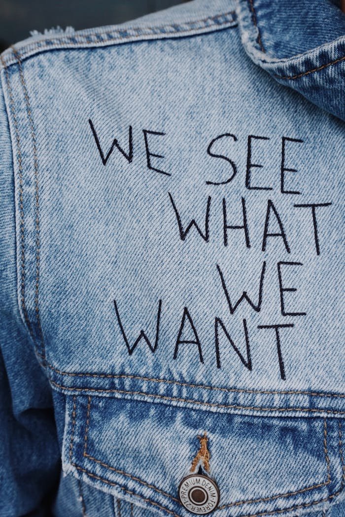 our-story Close-up of denim jacket featuring the phrase 'We See What We Want' embroidered.